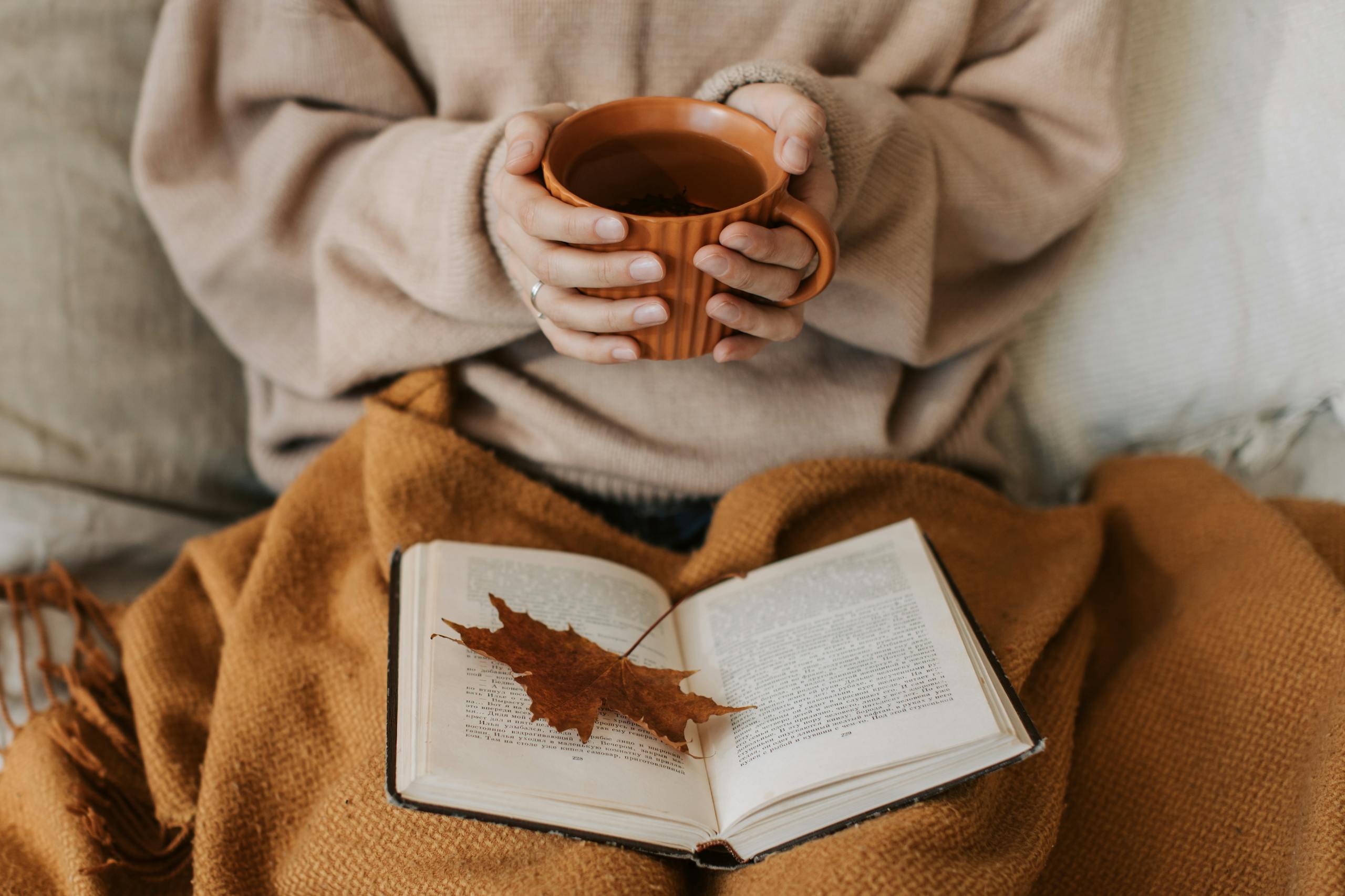 Une femme savoure un moment d'automne cocooning, tasse de thé en main au-dessus d'un livre et d'une feuille d'érable.