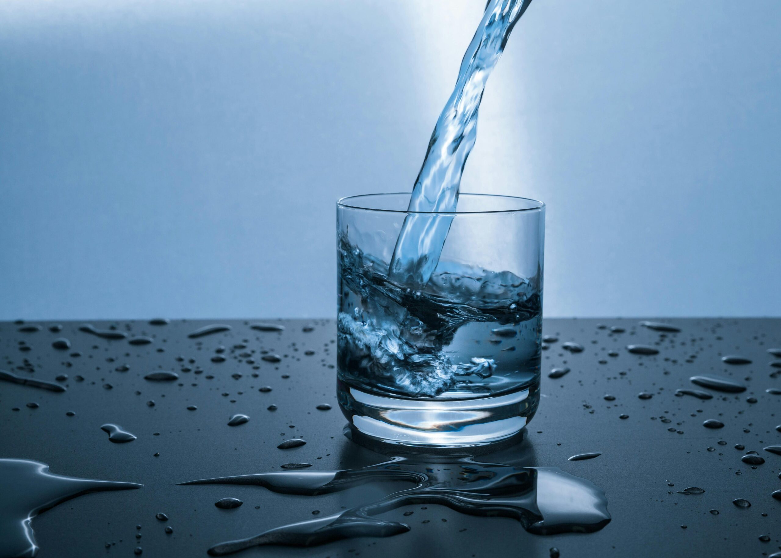 Un verre d'eau en train d'être rempli, posé sur une surface sombre parsemée de gouttelettes.